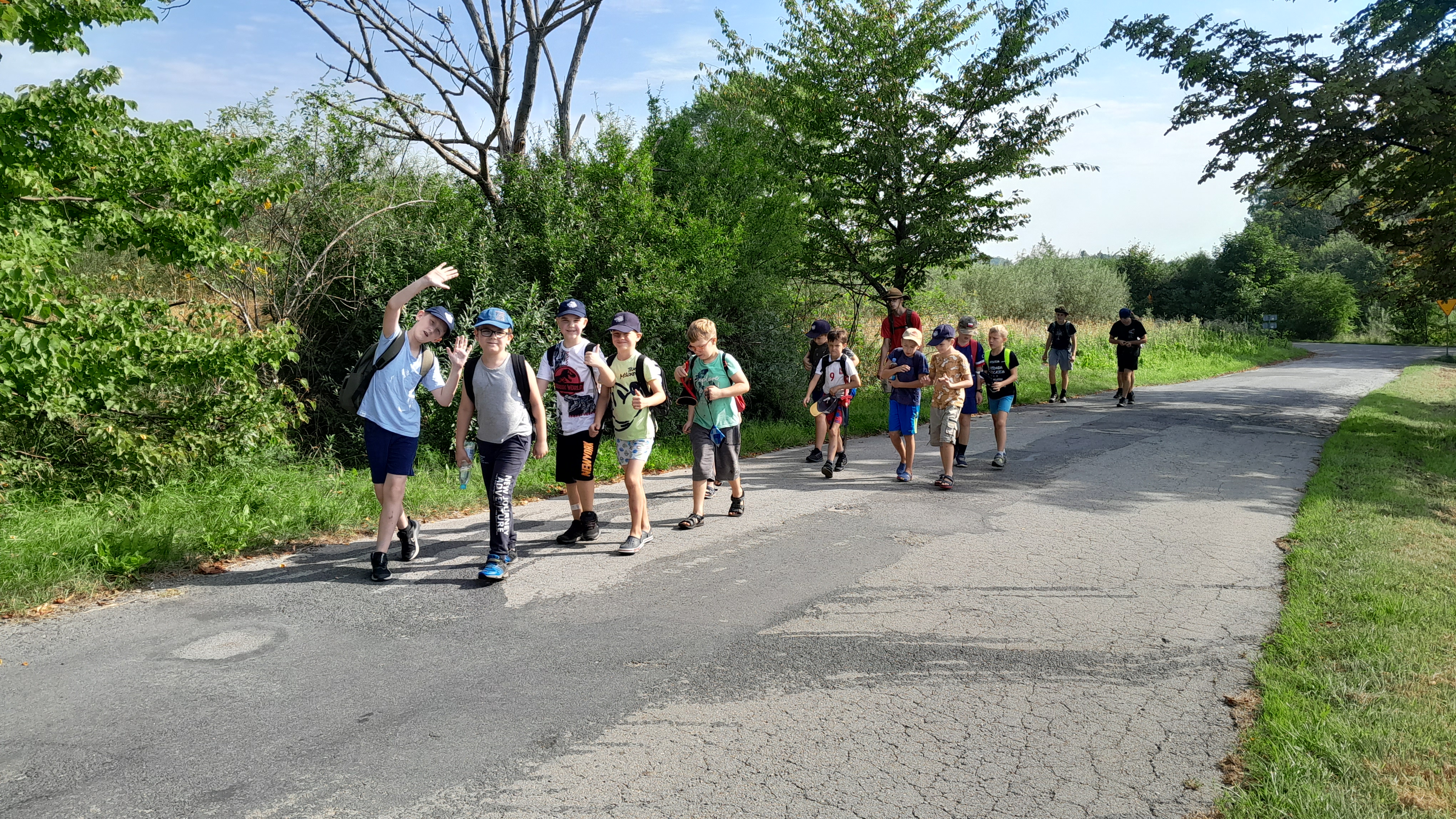 Na zdjęciu widać zuch&oacute;w na wędr&oacute;wce, znajdują się na drodze asfaltowej zuchy cieszą się i machają do kamery, idą w kolumnie, na głowach mają czapki zuchowe, z tyłu kolumny idą harcerze kt&oacute;rzy pilnują zuchy. Droga znajduje się w zalesionym terenie 