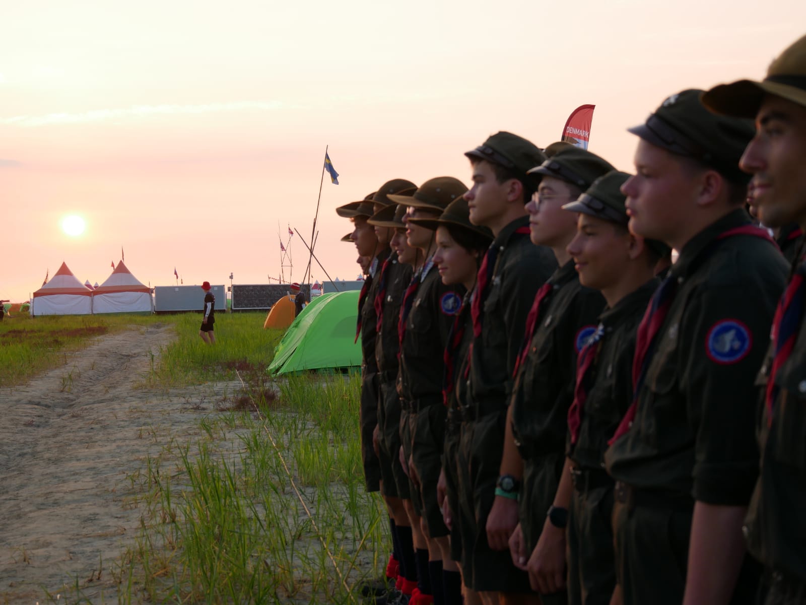 Harcerze stojący w szeregu aparat znajduje się po ich lewej stronie, harcerze mają na sobie mundury i barwy 5 Wodzisławskiej Drużyny Harcerzy Wilki (czerwony i granatowy), z tyłu widać jakiegoś przybłędę krzyczącego na swojego kolegę, widać r&oacute;wnież flagę Szwecji wywieszoną na wysokim maszcie, obraz jest w ciepłych kolorach spowodowane pięknym zachodem słońca nad koreańskim jamboree, widać r&oacute;wnież niewielkie namioty dwuosobowe w kt&oacute;rych śpią harcerze.